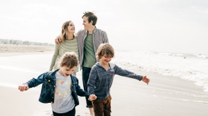 En familie på fire som går langs en strand. Foreldrene smiler og holder rundt hverandre, mens barna løper foran og holder hender, tydelig glade. Havet og bølgene skimtes i bakgrunnen under en lys og lett skyet himmel.