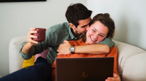 Et ungt par sitter i sofaen og smiler. Hun har en laptop i fanget og han holder rundt henne.