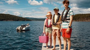 Tre unge personer som står på en brygge. En jente holder en rosa strandveske og har på en solhatt. To gutter står ved siden av henne. De har på seg redningsvester. En liten hvit båt er forankret på vannet i bakgrunnen.