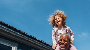 En liten jente sitter på skuldrene til pappaen sin og ler. Pappaen ler også. De er ute og det er fint vær.