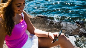 En ung kvinne sitter ved en innsjø og ser på mobilen sin. Hun smiler.