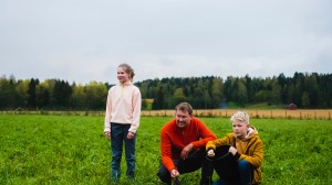 En mann og to barn ute på en grønn eng, med skog og åkrer i bakgrunnen