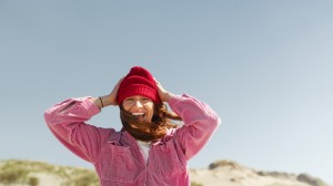 Smilende kvinne i lue og rosa jakke på sanddyner, med vind i håret.