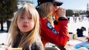 En jente med kakaobart sitter i skibakken og spiser appelsin. Mammaen i skiklær spiser matpakke