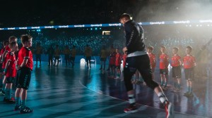 Landslagsmaskoter står på to rekker inne i en håndballhall mens en landslagsspiller løper ut på banen mellom dem