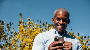 En smilende mann ser på mobilen sin, stående foran blomstrende busker under en blå himmel.