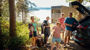 En familie med to voksne og tre barn står foran bilen og hytta si med bagasje.