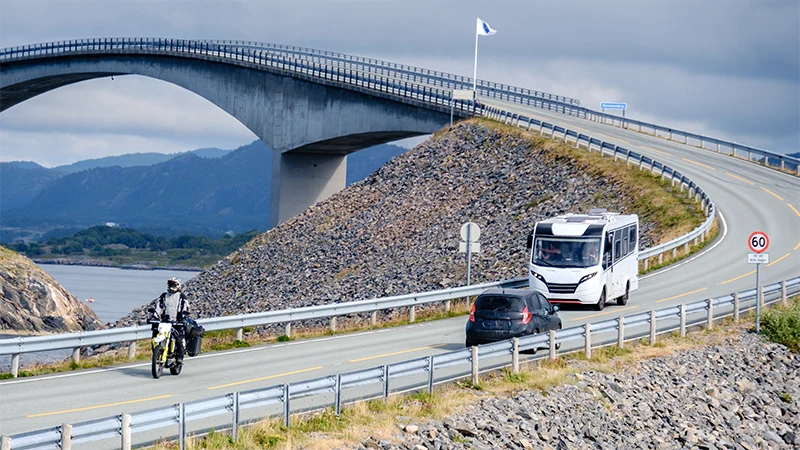 En motorsykkel, en personbil og en bobil kjører på veien. I bakgrunnen er en stor bro.