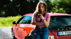 En ung kvinne står foran en bil med øvelseskjøringsskilt. Hun sjekker Øvelseskjøringappen på mobilen sin. 