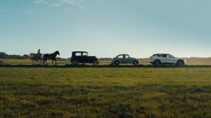 Hest og kjerre, gamle biler og til slutt en moderne bil kjører på rekke på en landevei.