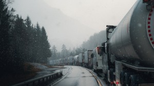 Kø av lastebilder med tilhengere på vinterføre langs svingete vei.