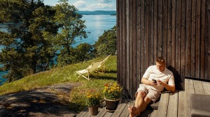En mann sitter på trappen utenfor hytta si og ser på mobilen. Trær og hav i bakgrunnen.
