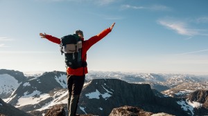 En kvinne med ryggsekk står på fjelltopp med armene løftet, omgitt av snødekte fjell.