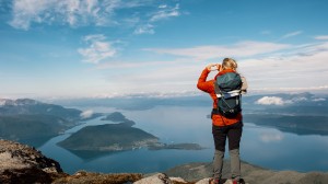 En kvinne står på toppen av et fjell og lager et hjerte med hendene sine, mens hun ser utover fjorden.