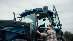 Blå traktor med hvite hjul står på et jorde. En mann sitter i førerhuset, mens en kvinne og et barn står ved forhjulet