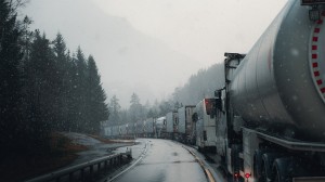 Kø av lastebilder med tilhengere på vinterføre langs svingete vei.