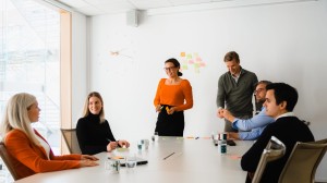 Six colleagues are gathered around a large table in a meeting room. There are post-it notes on the walls, and they're smiling as they listen to what Ane is saying.