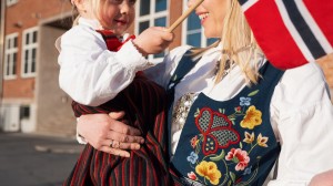 Mor har løftet opp datteren sin og holder henne inntil seg. De har på seg bunader, vifter med det norske flagget og smiler.