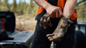 Tre personer står ved en grind og ser utover et jorde, mens en svart og hvit hund går i nærheten.