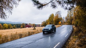 En bil kjører på en våt landevei gjennom et høstlig landskap med gule trær og en rød låve i bakgrunnen.