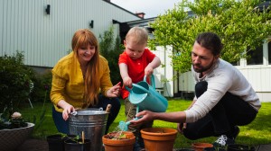 Familie på to voksne og to små barn sitter på huk i hagen og planter grønnsaker og vanner de.