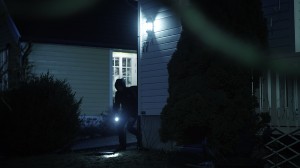 En person med huvtröja och ficklampa kommer smygandes runt knuten av ett hus i mörkret