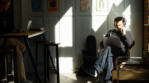 En mann sitter på en kafe' med en laptop på fanget og snakker i telefonen.