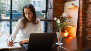 En ung kvinne sitter på en kafé med en bærbar PC og skriver notater.