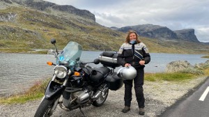Karen har parkert motorsykkelen ved et vann langs veien. Hun har tatt av hjelmen og smiler til kameraet med fjell i bakgrunnen.