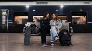 Et ungt par sitter på en benk og venter på toget. De smiler og ser på hverandre.