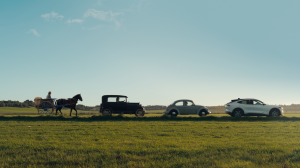 Hest og kjerre, gamle biler og til slutt en moderne bil kjører på rekke på en landevei.