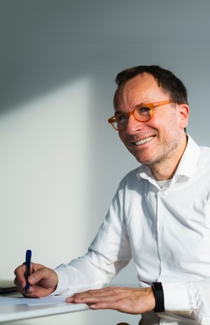 Christian sits by a desk, wearing a white shirt and orange glasses, smiling as he draws with a pen on paper.