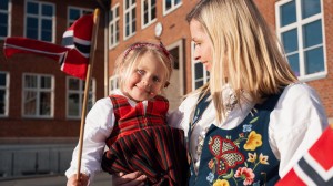 Mor har løftet opp og holder datteren sin inntil seg. De har på seg bunader, vifter med det norske flagget og smiler.